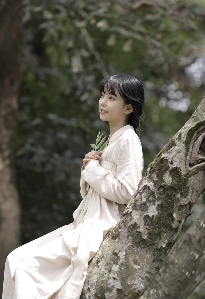 一名身穿白色长裙的女子靠在森林中的树旁。
