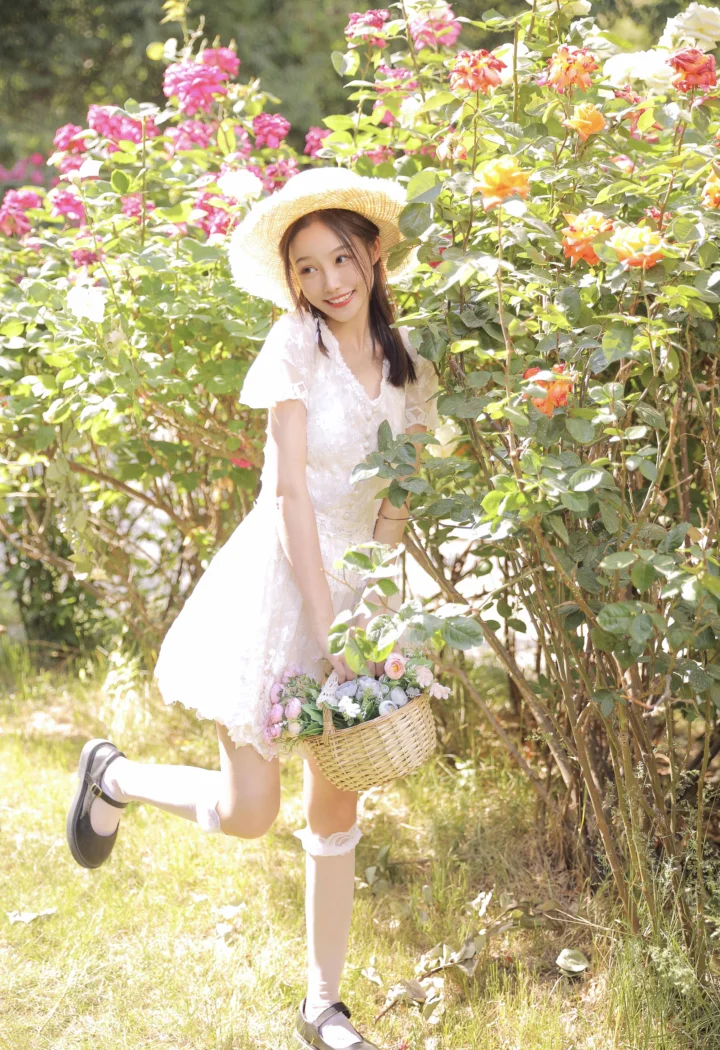 麦わら帽子と白いワンピースを着た若い女性が、咲き誇るバラの中で花かごを持って立っています。