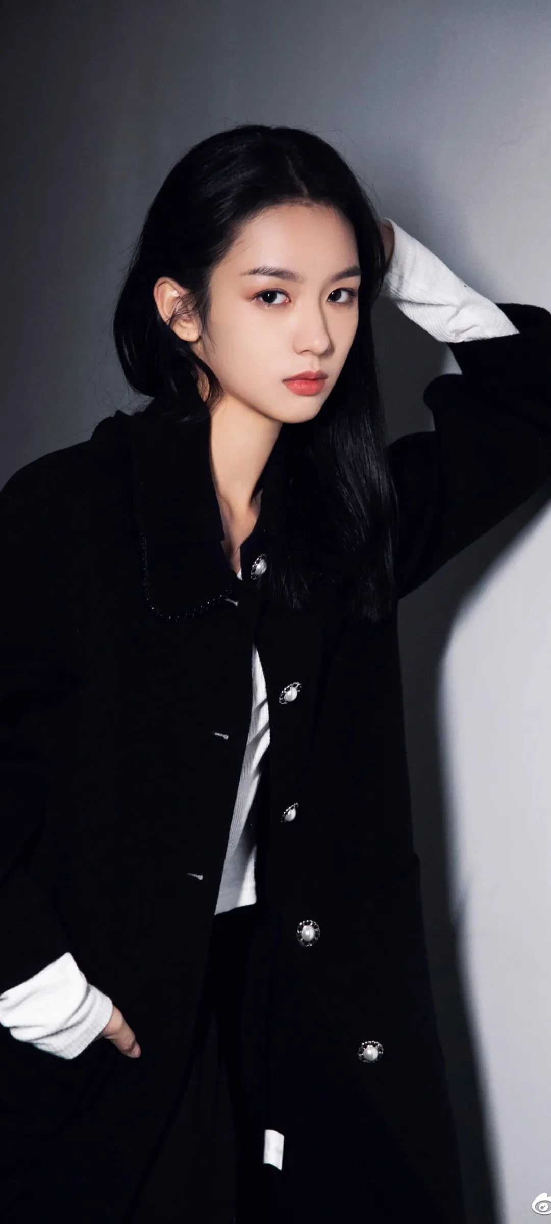 긴 검은 머리의 여성이 회색 배경 앞에서 검은 코트를 입고 포즈를 취하고 있다.