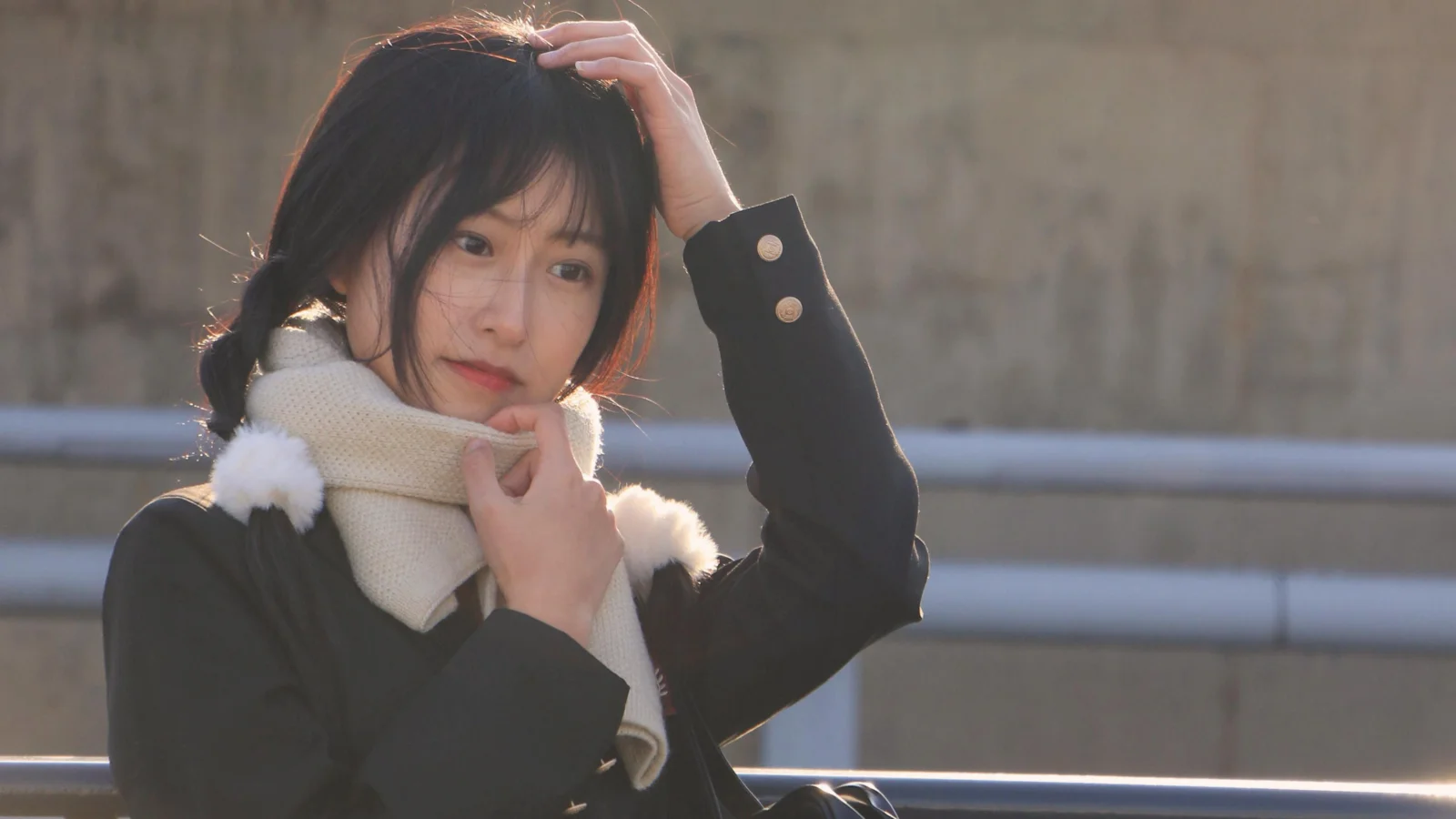 一名戴着围巾的女子站在户外栏杆旁，阳光从侧后方照亮她的头发。