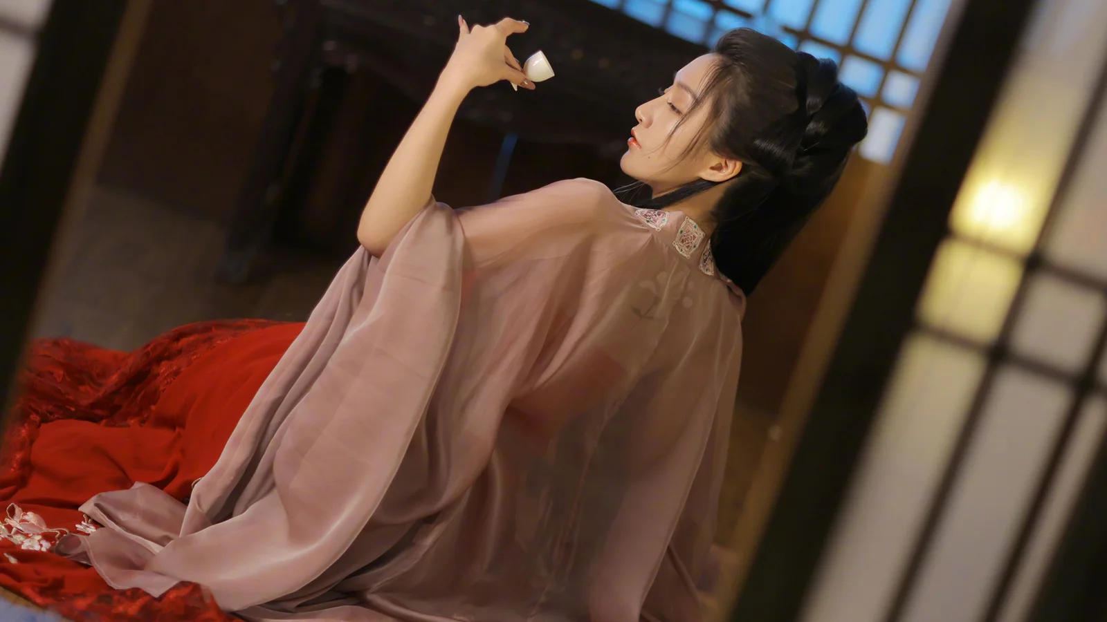 一名身穿浅色宽袖服饰的女子坐在红色织物旁，手持小杯，背景为室内木质窗格与暖色灯光。