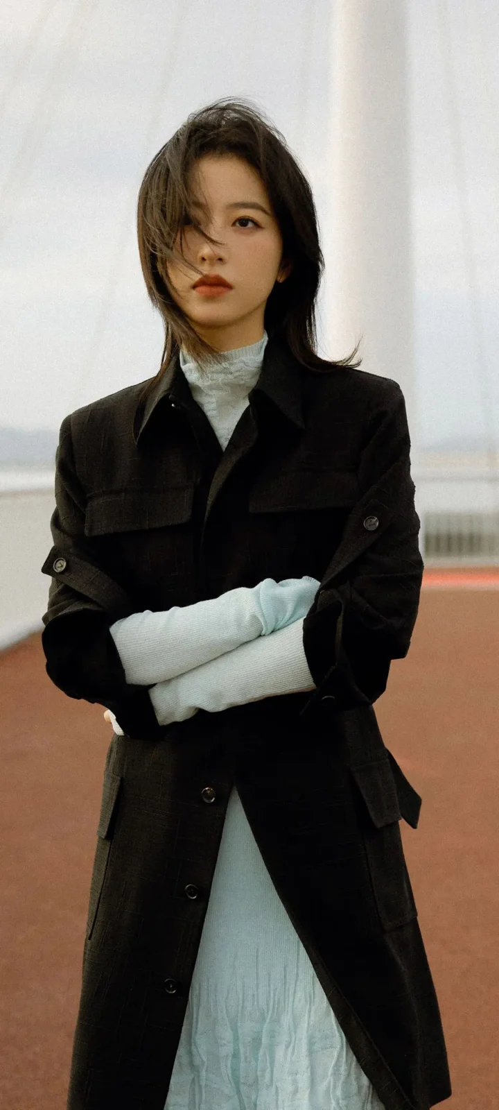 검은 코트와 밝은 색상의 옷을 입은 성인 여성이 실내 배경 앞에서 카메라를 바라보고 있다.