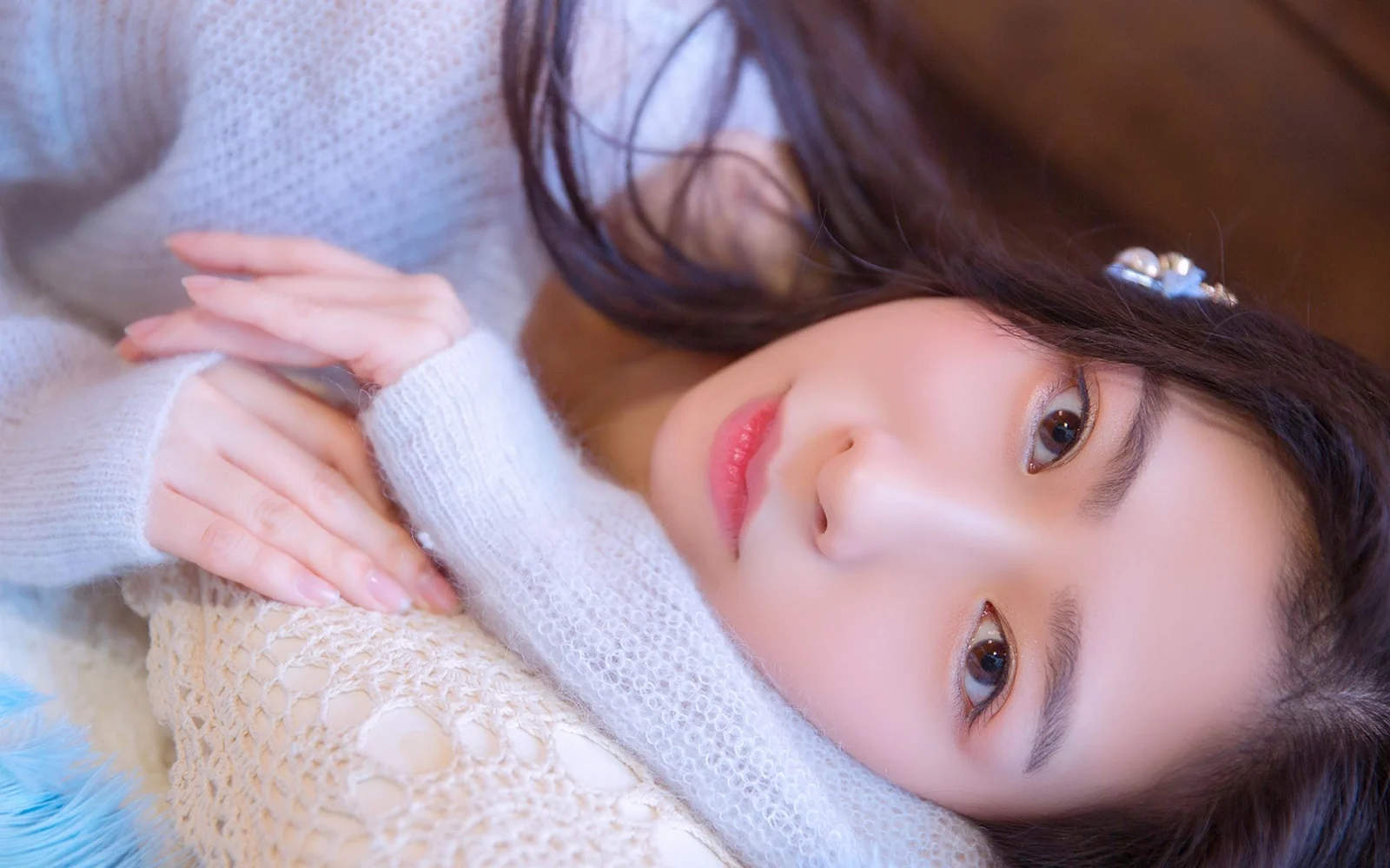 淡い色のニットを着た女性が柔らかな編み物の上に横向きに寝そべり、カメラを見つめています。