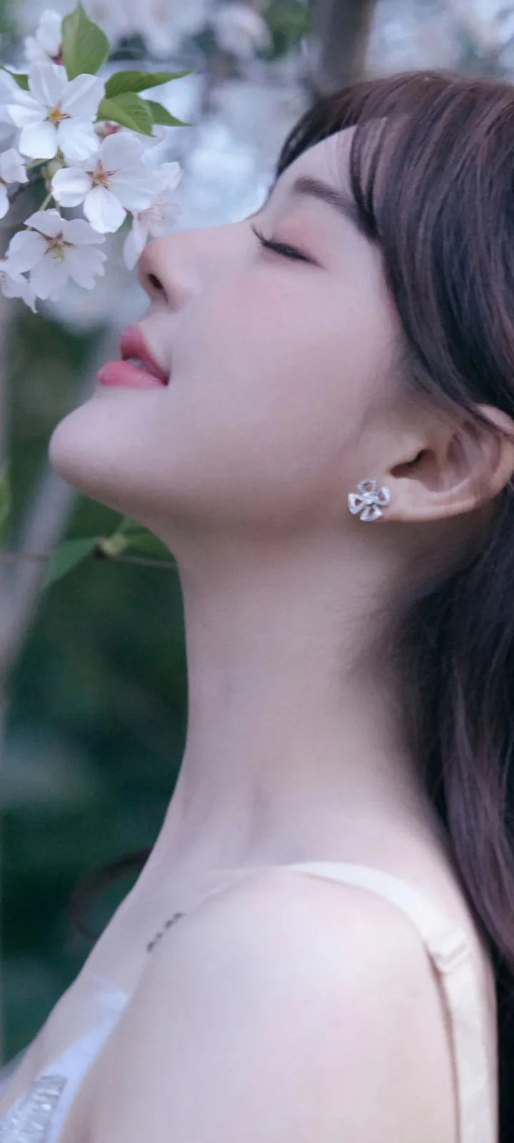 一位成年女性闭眼侧脸靠近白色花朵，背景柔和虚化。