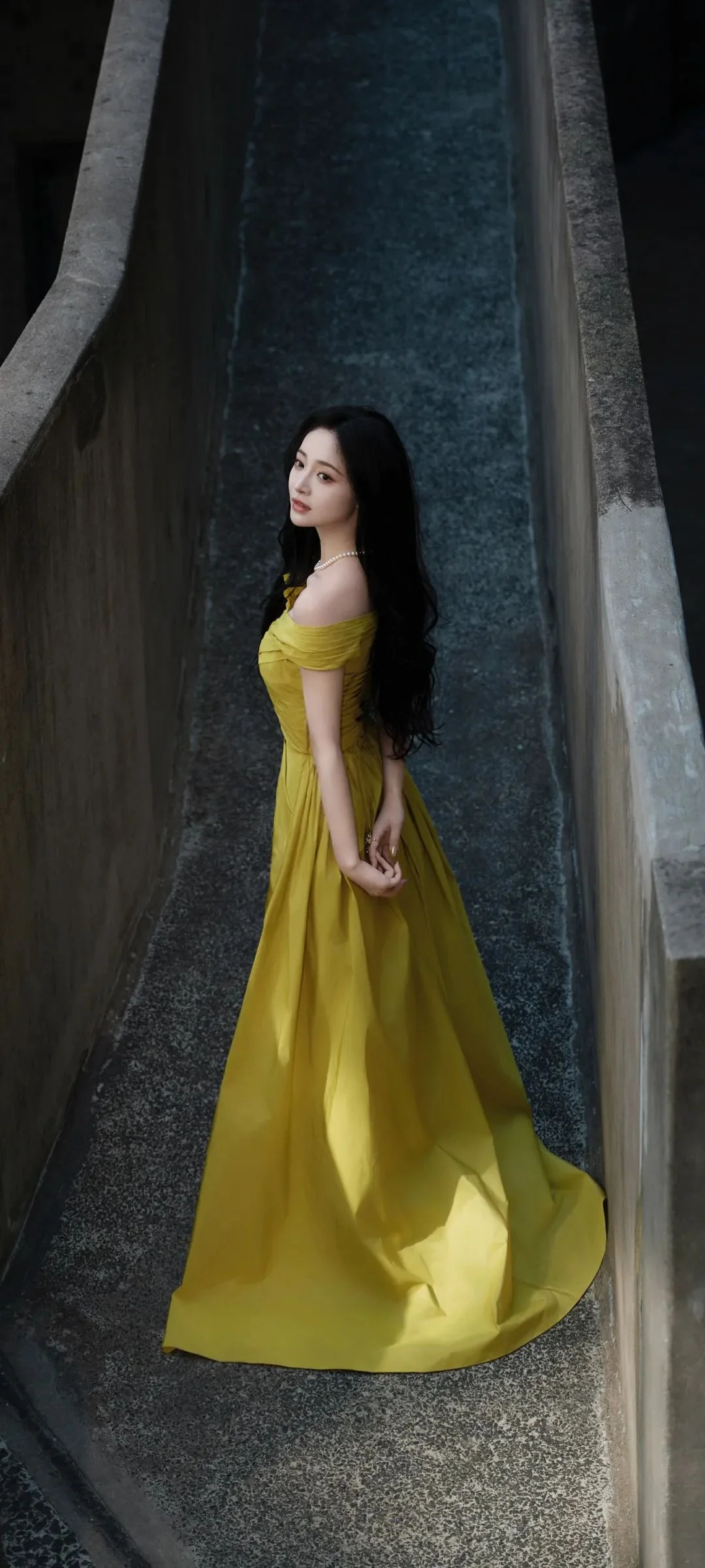 노란색 롱드레스를 입은 여성이 좁은 석조 건축 통로에 서 있다.