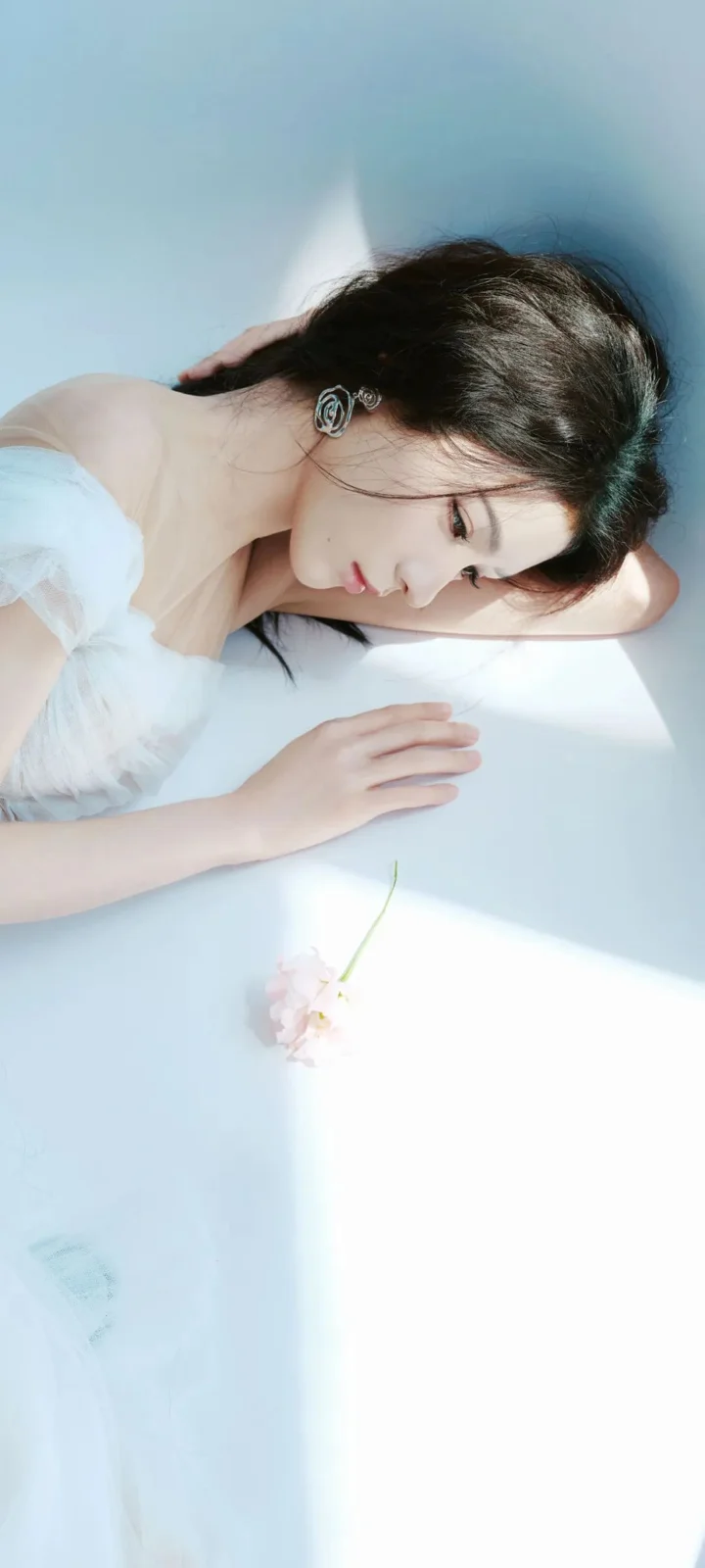 一名黑发女子身穿白色衣服侧卧在浅色表面上，柔和的蓝色光线洒在她身上。