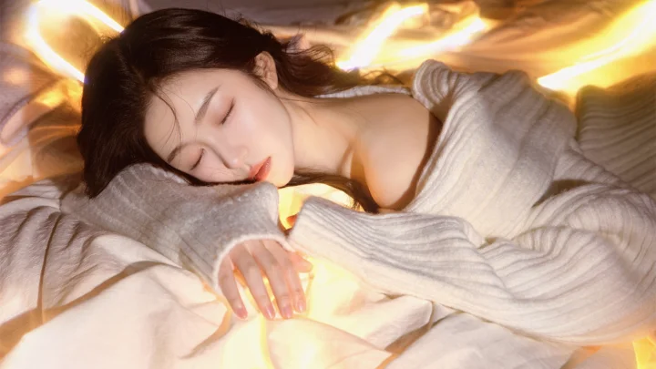 一名女子穿着浅色针织衫，在温暖的灯光下躺在床上熟睡。