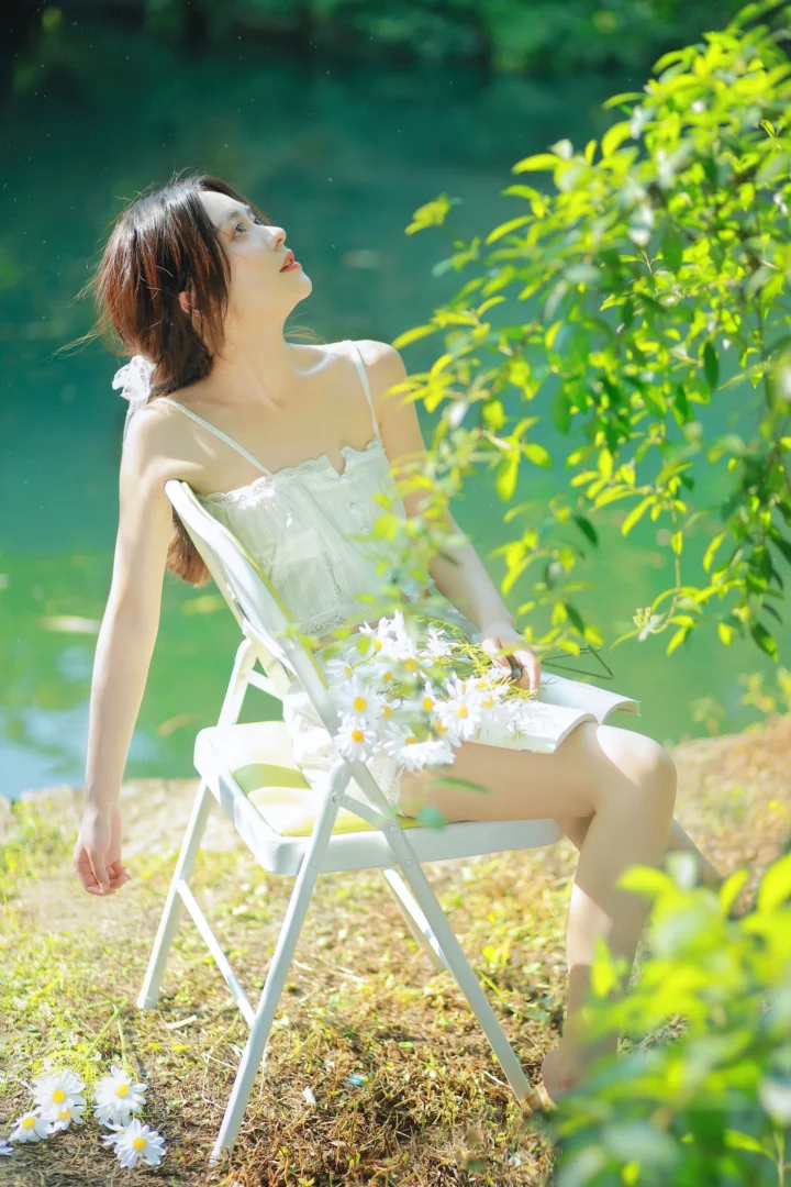 흰색 원피스를 입은 여성이 호숫가 접이식 의자에 앉아 밝은 자연광 속에서 데이지를 안고 있다.