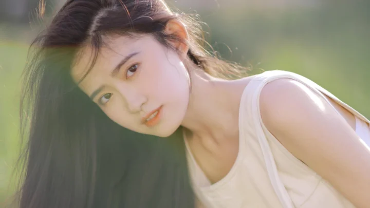 一名长发女子在柔和阳光下靠近镜头看向前方，背景为虚化的绿色户外景色。