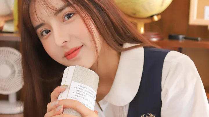 一位长发女子手持书本，在室内暖色背景前看向镜头。