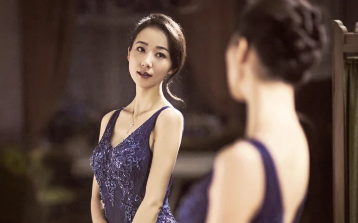 一名成年女子穿着蓝色连衣裙站在镜子前，画面中可见她的倒影。