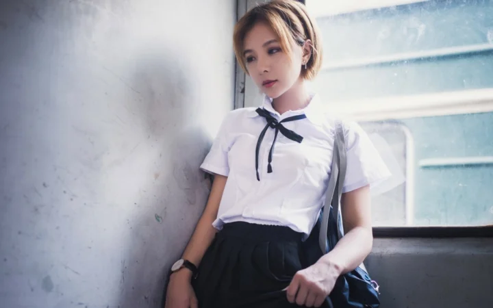 一名身穿白色上衣和深色裙子的女性站在明亮窗边。