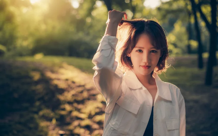 흰 셔츠를 입은 성인 여성이 햇살이 비치는 공원에서 한 손을 머리에 올리고 서 있습니다.