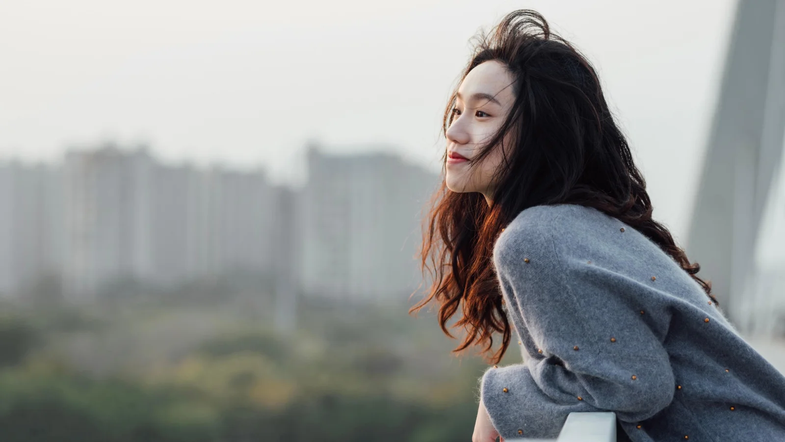 一名长发女性穿着灰色毛衣站在户外，身后是虚化的城市建筑。
