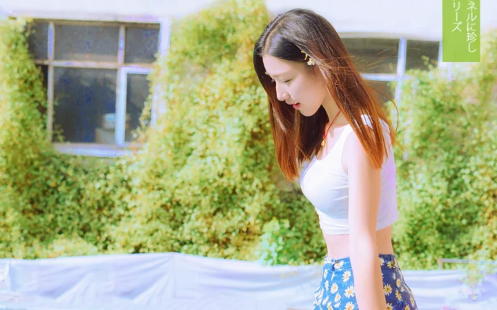 一名成年女子站在绿植墙前，穿着白色上衣和花卉裙子，低头望向一侧。