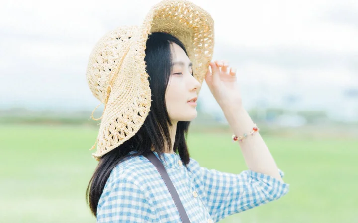 一名戴草帽、穿蓝白格纹衬衫的女子站在绿色田野中。