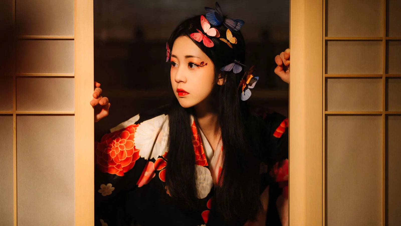 나비 머리 장식과 긴 검은 머리를 한 여성이 나무문 사이에 서서 옆을 바라보고 있다.
