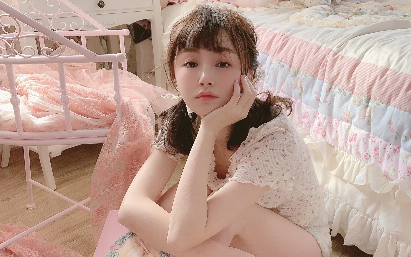 짧은 머리의 여성이 분홍색 침실에서 침대 옆에 앉아 손으로 뺨을 받치고 카메라를 바라보고 있다.
