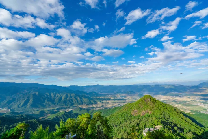 蓝天白云下，层叠山脉环绕着绿色山谷与远处的田野。
