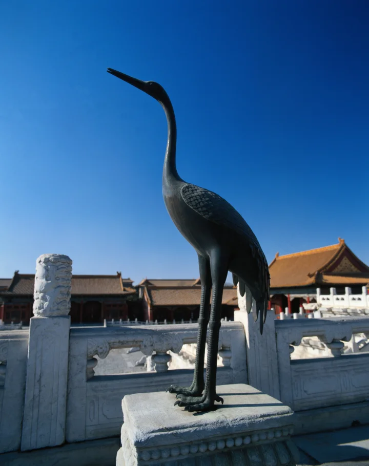 一只青铜鹤雕像矗立在宫殿建筑与湛蓝天空前方。