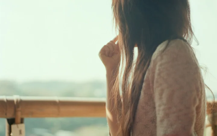 長い髪の女性が明るい屋外の景色に向かってベランダに立っている。