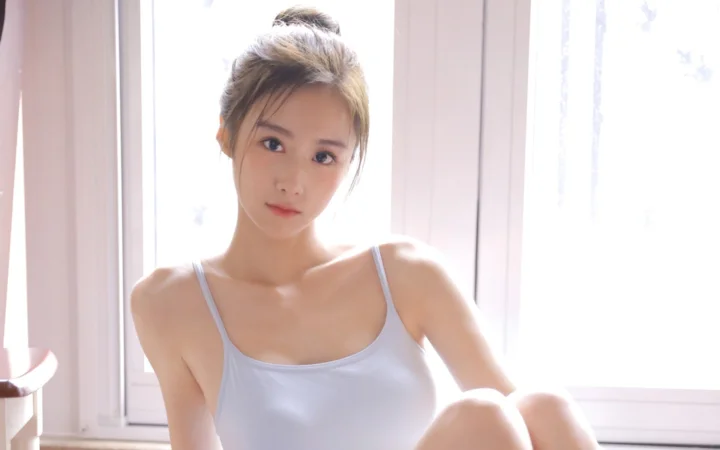 一位成年女性坐在明亮窗边，身穿白色细肩带上衣，面向镜头微笑。