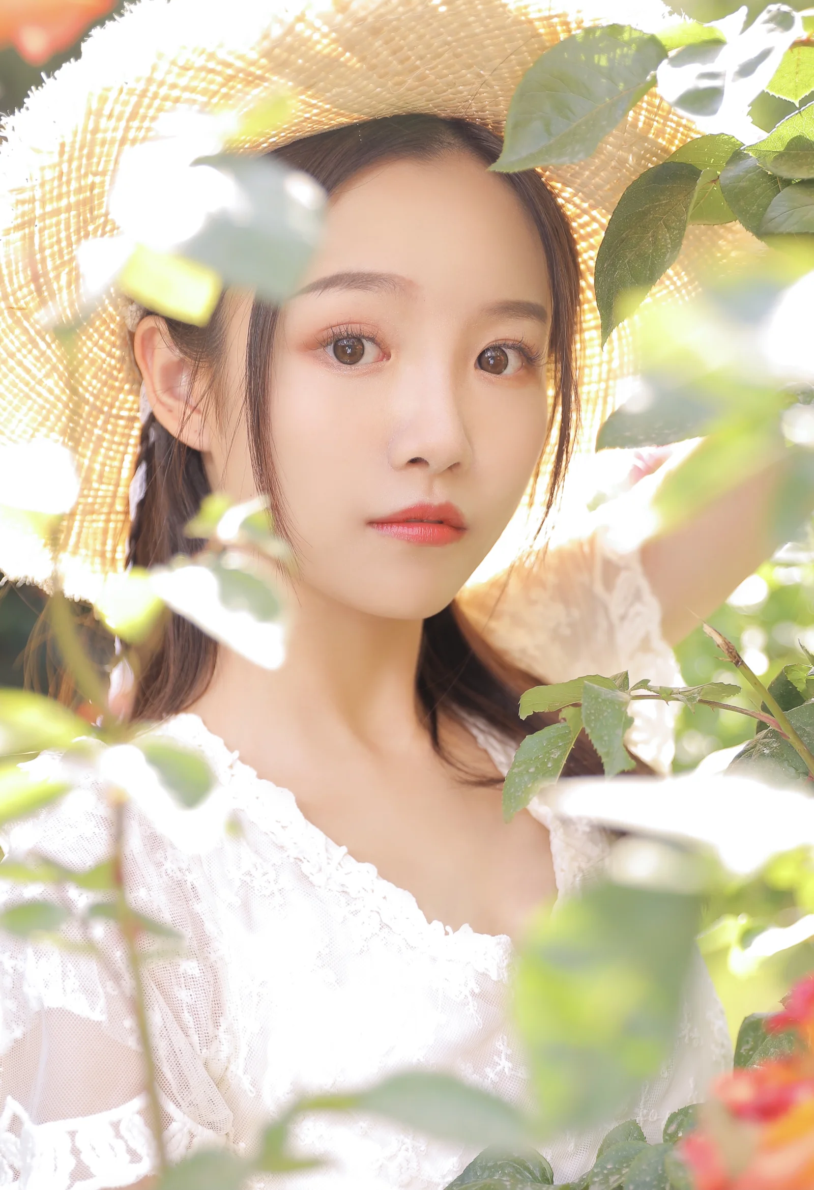 麦わら帽子をかぶった若い女性が、陽光と緑の葉に囲まれてカメラを見つめています。