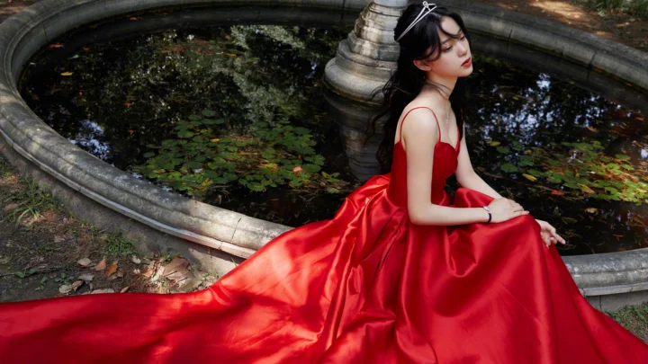 一名身穿红色长裙的女子坐在户外喷泉池旁。