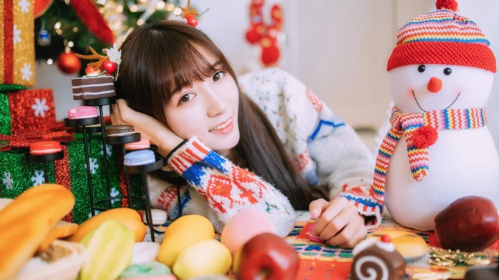 女性がクリスマスの飾りやカラフルな贈り物、編みぐるみの雪だるまのそばでカメラに微笑みかけています。