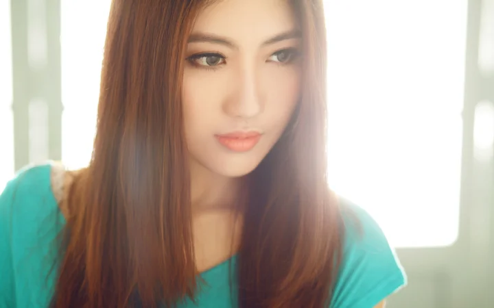 긴 머리의 여성이 청록색 상의를 입고 밝은 창가에 서 있습니다.