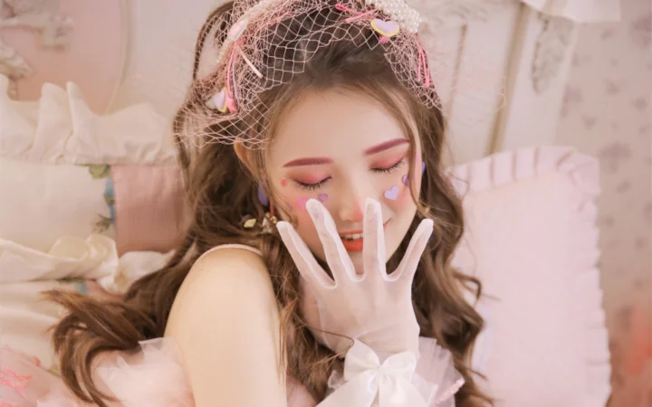 흰색 긴 장갑을 끼고 컬러 메이크업과 하트 스티커를 한 여성이 분홍빛 실내에 앉아 있다.
