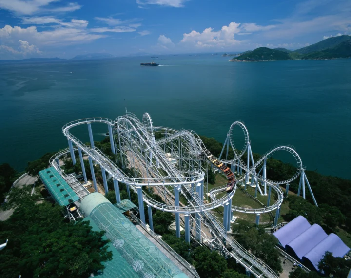白色过山车盘旋在临海游乐园中，远处是湛蓝海湾、岛屿和群山。