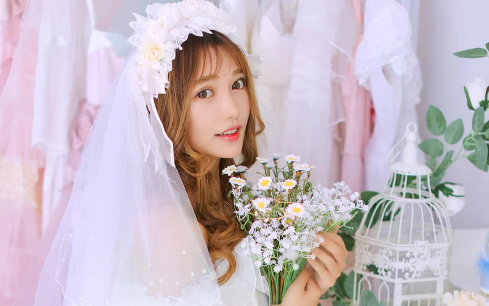 白いベールを着けて小さな花束を持つ女性が、明るく柔らかく装飾された室内でカメラを振り返っている。