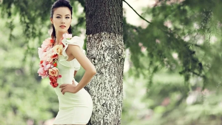 一名成年女性身穿白色连衣裙，手持花束站在树旁。