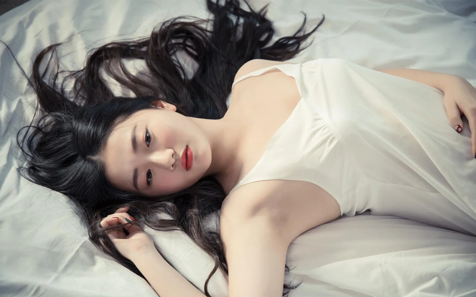 검은 머리의 여성이 흰색 민소매 옷을 입고 흰 침대에 누워 있습니다.