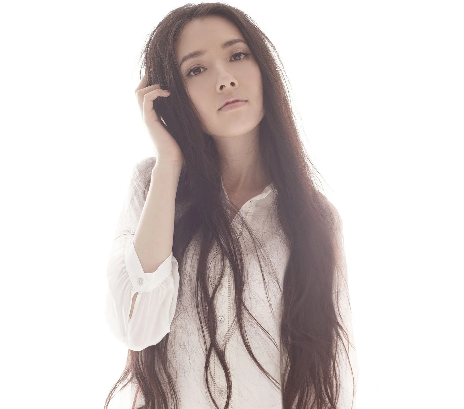 긴 머리의 여성이 흰 셔츠를 입고 밝은 배경 앞에 서 있다.