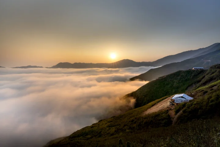 晨光照耀群山与翻涌的云海，山坡上可见一座小屋。