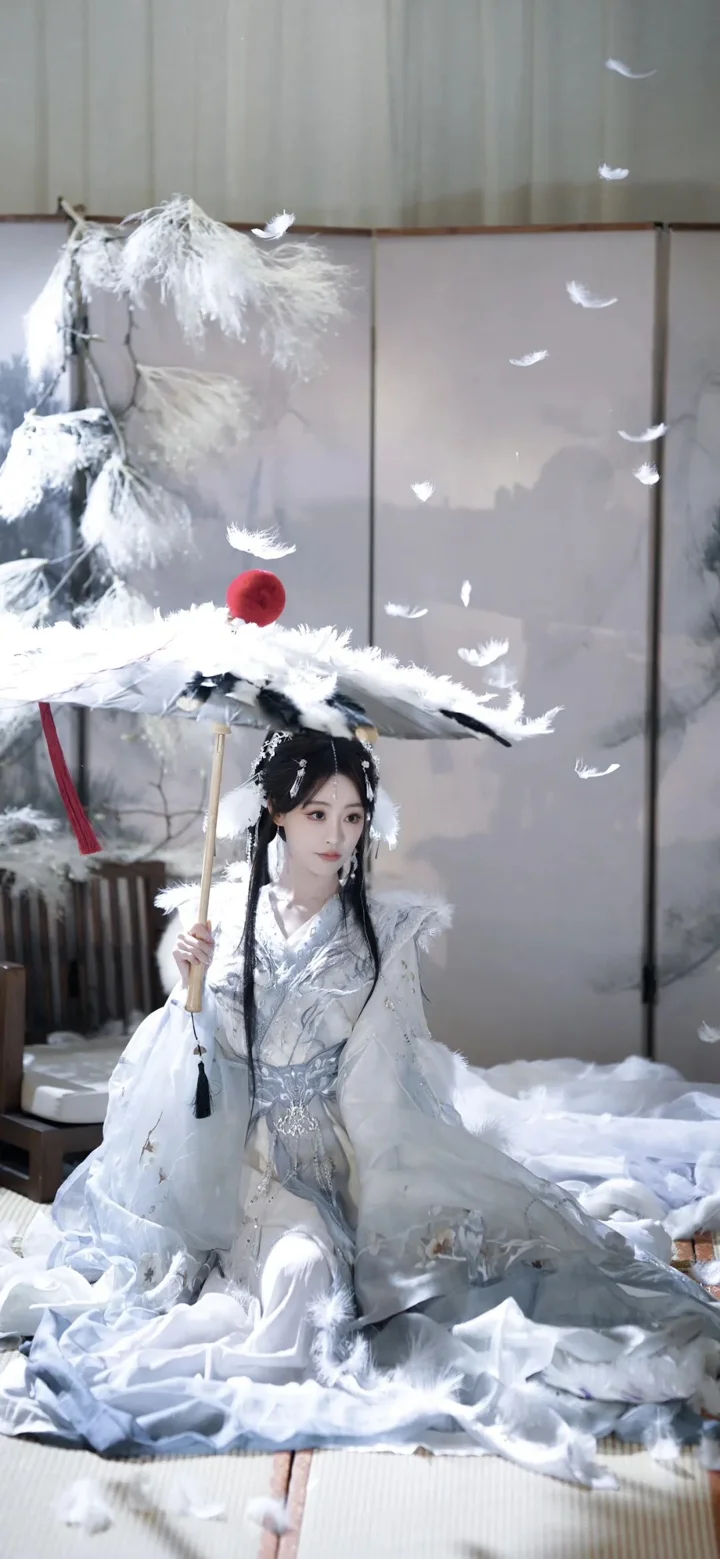 一名身穿白色古装的女子坐在室内，手持装饰着羽毛的伞，周围散落着白色羽毛。