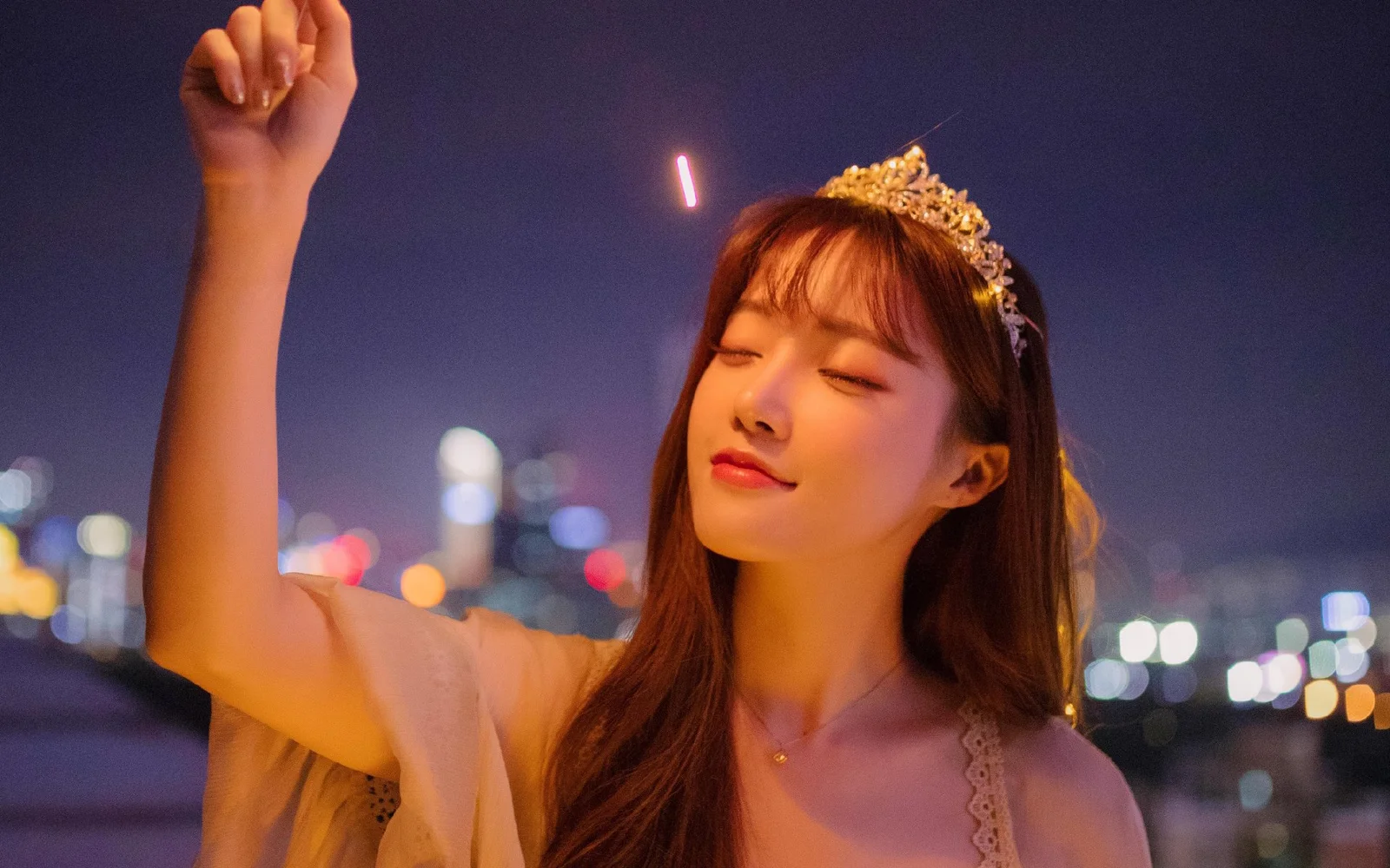 一名戴着皇冠的女性在夜晚城市灯光前闭眼举起手臂。