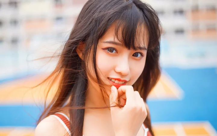 검은 머리의 여성이 손가락을 턱 가까이에 대고 부드럽게 흐려진 파란색과 주황색 배경 앞에서 카메라를 향해 미소 짓고 있다.