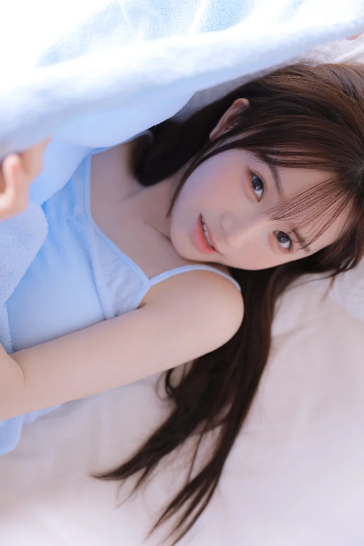 一名棕色长发女性躺在浅色床上，面向镜头微笑。