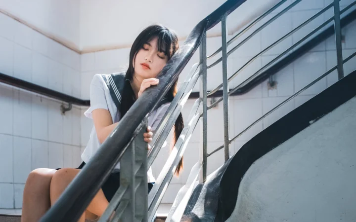 一名成年女性坐在室内楼梯旁，阳光从一侧照亮她和金属扶手。