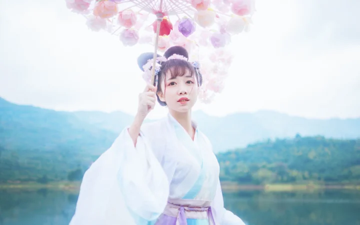 一名身穿白色传统服饰的女子撑着花伞站在湖畔，远处是群山。
