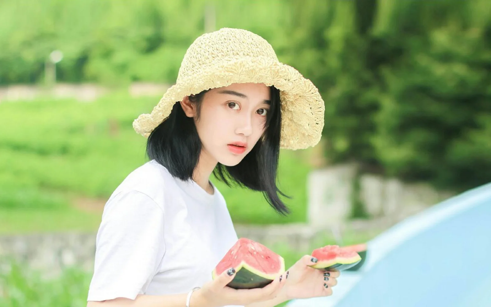 밀짚모자를 쓴 여성이 푸른 야외 배경 앞에서 수박 조각을 들고 있습니다.