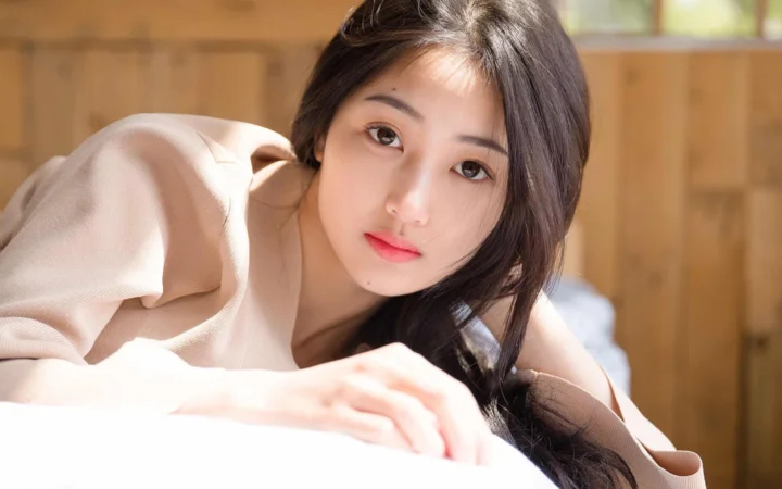 一名长发女性穿着浅色上衣，在温暖木质室内靠近镜头望向前方。