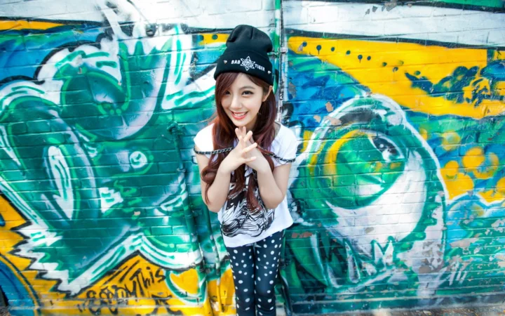 帽子をかぶった女性が、鮮やかなグラフィティの壁の前で微笑んでいる。