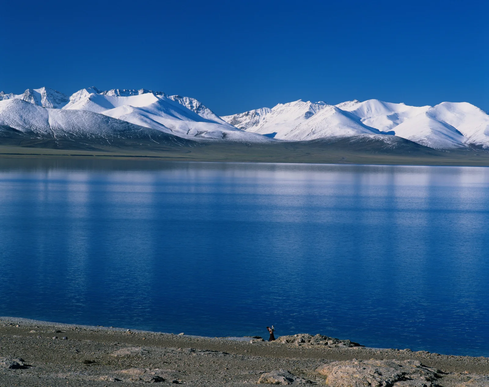蓝色湖泊对岸绵延着覆雪山脉，前景是荒凉的岩石湖岸。