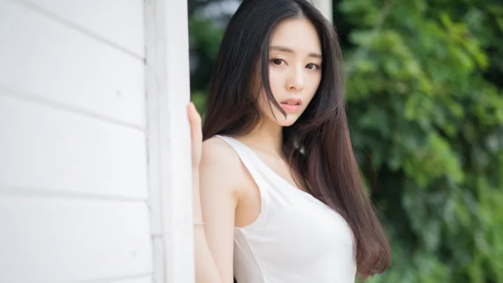 一名长发女子穿着白色上衣站在窗边，背景是绿色植物。