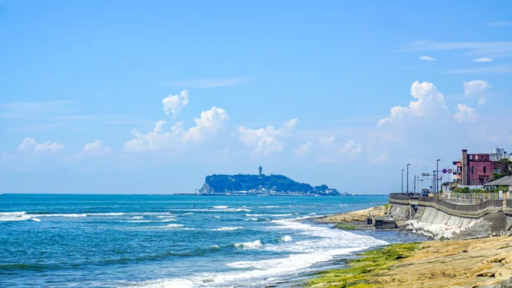 湛蓝海水拍打着海岸，远处的小岛、白云与临海建筑共同构成明亮的海滨景色。