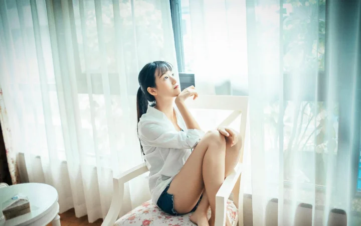 一名成年女子穿着白色上衣坐在窗边的白色椅子上，柔和的自然光透过窗帘洒入室内。
